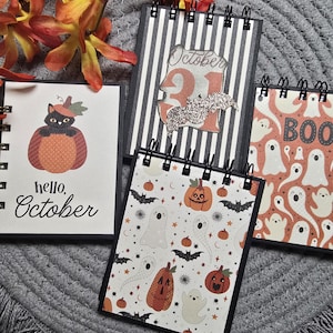 Può includere: Quattro quaderni a spirale con copertine a tema Halloween. Uno mostra un gatto nero in una zucca con il testo 'hello, October'. Un altro ha strisce bianche e nere con 'October 31' e un pipistrello glitterato. Un terzo mostra fantasmi e la scritta 'BOO'.