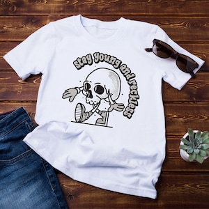 Op de afbeelding: Wit T-shirt met een zwart-witte illustratie van een cartoon schedel met schoenen en handschoenen. De tekst "Stay young and reckless" staat in een boog boven de schedel.