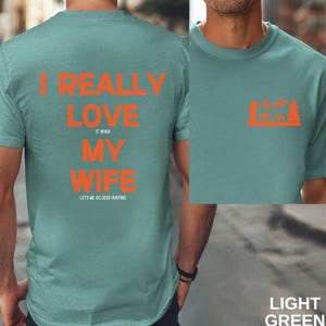 K&ouml;nnte beinhalten: Ein hellgr&uuml;nes T-Shirt mit dem Text "I really love my wife it when lets me go deer hunting" in Orange. Auf der R&uuml;ckseite des Shirts ist eine Silhouette eines Hirsches in einem Wald zu sehen.