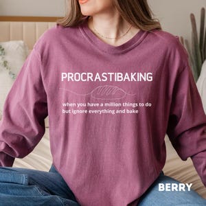 May include: Berry-colored sweatshirt with the text "PROCRASTIBAKING" above a bread loaf illustration. The text below reads "when you have a million things to do but ignore everything and bake." The word "BERRY" is printed at the bottom.
