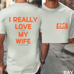 K&ouml;nnte beinhalten: Wei&szlig;es T-Shirt mit orangefarbenem Text, der "Ich liebe meine Frau wirklich, wenn sie mich zum Hirschjagen l&auml;sst" lautet. Die R&uuml;ckseite des Shirts hat eine kleine orangefarbene Grafik einer Bergszene mit zwei Hirschen.