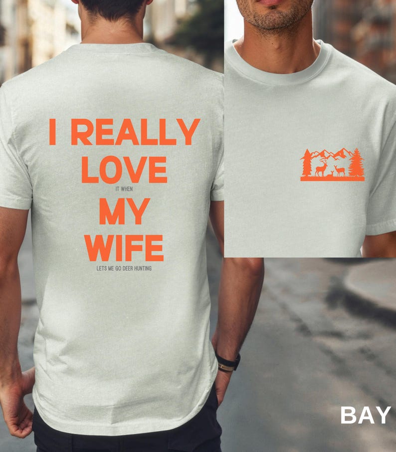 K&ouml;nnte beinhalten: Wei&szlig;es T-Shirt mit orangefarbenem Text, der "Ich liebe meine Frau wirklich, wenn sie mich zum Hirschjagen l&auml;sst" lautet. Die R&uuml;ckseite des Shirts hat eine kleine orangefarbene Grafik einer Bergszene mit zwei Hirschen.