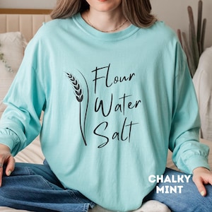 Może przedstawiać: Jasno błękitny t-shirt z długim rękawem z napisem "Flour Water Salt" i grafiką kłosa pszenicy. Koszulka jest oznaczona jako "Chalky Mint".