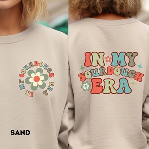 May include: Sand-colored sweatshirt with a floral design and the text "IN MY SOURDOUGH ERA" on the back. The front features a flower graphic with the words "IN MY SOURDOUGH ERA" around it. The word "SAND" is printed at the bottom.
