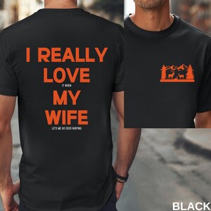 K&ouml;nnte beinhalten: Schwarzes T-Shirt mit orangefarbenem Text, der "Ich liebe meine Frau wirklich, wenn sie mich zum Hirschjagen l&auml;sst" lautet. Die R&uuml;ckseite des Shirts hat eine kleine orangefarbene Grafik einer Bergszene mit zwei Hirschen.