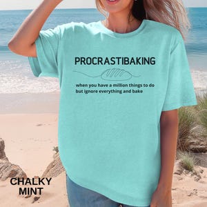 May include: A light teal t-shirt with the words "PROCRASTIBAKING" above a bread loaf illustration. Below the illustration, the text reads "when you have a million things to do but ignore everything and bake." The shirt is labeled "CHALKY MINT."