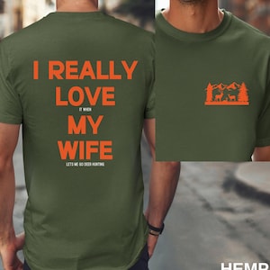 K&ouml;nnte beinhalten: Ein gr&uuml;nes T-Shirt mit dem Text "I really love my wife it when lets me go deer hunting" auf der R&uuml;ckseite. Die Vorderseite des Shirts zeigt eine Grafik mit einer Waldszene mit Hirschen.