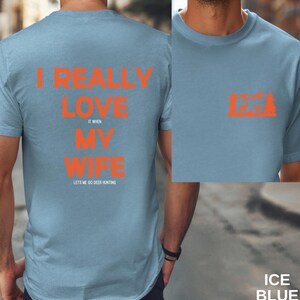 K&ouml;nnte beinhalten: Ein hellblaues T-Shirt mit dem Text "I really love my wife it when lets me go deer hunting" in orange gedruckt. Die R&uuml;ckseite des Shirts zeigt eine kleine orangefarbene Grafik eines Hirsches in einem Wald.