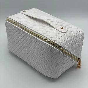 May include: White woven cosmetic bag with a gold zipper and a gold handle. The bag has a textured woven design.