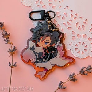 May include: A clear acrylic charm depicting a cartoon character with blue eyes and dark hair, mounted on a white and pink winged creature. The charm has a black clasp and a gold-coloured wing charm. The background is pink with lavender sprigs.