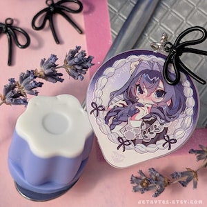 May include: A lavender-colored, flower-shaped soap dish with a white top, next to a round anime-style character charm. The charm features a girl with purple hair and a white lace border. Black bow-shaped hair clips and dried lavender sprigs are also present.