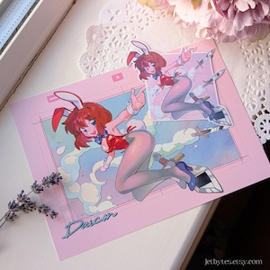 Puede incluir: Una impresión rosa con un personaje de dibujos animados con un disfraz de conejito, cabello rojo, un atuendo rojo y blanco y zapatos azules. La impresión incluye el nombre "Daisen" en cursiva. También hay una pegatina más pequeña del personaje.