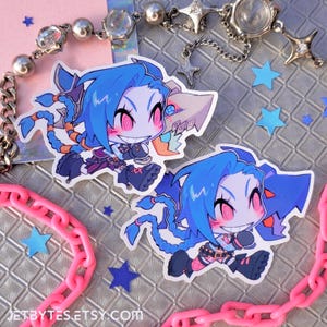 May include: Two cartoon stickers of Jinx from League of Legends. The stickers are on a silver background with pink chain links and blue stars. Jinx is a blue-haired character with red eyes and a mischievous grin. She is wearing a black and white outfit and has a pink and white weapon.