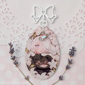 Peut inclure: Un porte-clés ovale de style anime avec un personnage entouré de fleurs blanches. Le personnage a les cheveux roses et porte une tenue noire. Le porte-clés a un dessus en forme de nœud blanc et un petit charme en forme de cœur. Le fond est rose.