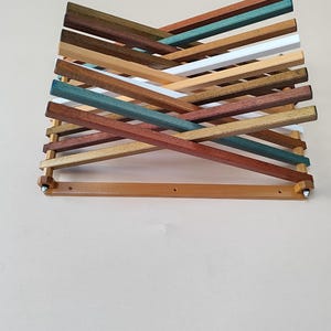 Könnte beinhalten: Ein Wandkunstwerk aus Holz mit geometrischem Design. Das Kunstwerk besteht aus mehreren Holzlatten in verschiedenen Farben, darunter Braun, Grün, Weiß und Türkis, die in einem sich kreuzenden Muster angeordnet sind. Das Werk ist an einer hellen Wand befestigt.