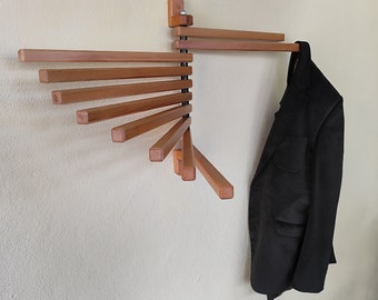 May include: A wooden wall-mounted coat rack with a folding design. The rack is made of natural wood and has a dark brown finish. A black blazer is hanging on the rack.