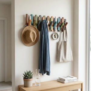 May include: A wall-mounted coat rack featuring multiple vertical wooden pegs in various shades of blue, green, brown, and white. The rack holds a straw hat, a blue knit sweater, a small circular tray with keys, and a beige canvas tote bag.