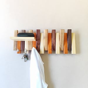 Könnte beinhalten: Wandmontierter Kleiderständer aus Holz mit einem Regal und Schlüsselhaken. Der Kleiderständer besteht aus verschiedenen Holzstücken in unterschiedlichen Farben und hat ein schwarzes Metallregal.