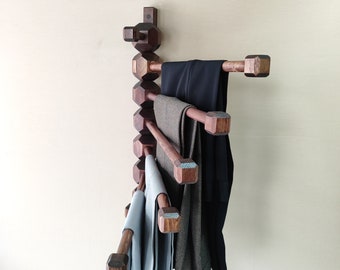May include: A wooden wall-mounted clothes rack with five arms. The rack is made of dark brown wood and has a geometric design. The rack is holding a variety of clothing, including a pair of black pants, a pair of brown pants, and a light blue shirt.