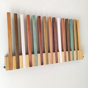 May include: A wooden wall-mounted coat rack with a series of vertical wooden slats in various colors, including brown, white, and teal. The rack is mounted on a white wall and has a rectangular base.