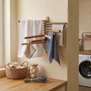 Könnte beinhalten: Ein wandmontierter Handtuchhalter aus Holz mit mehreren schwenkbaren Armen, an denen ein weißes Handtuch, ein beiges Tuch und ein blaues strukturiertes Handtuch hängen. Ein geflochtener Korb, ein Glas mit weißen Kugeln und Lavendel.