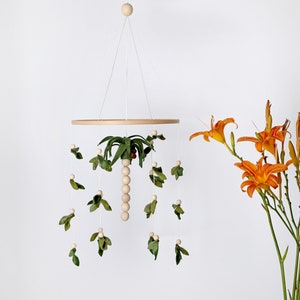 May include: A wooden baby mobile with a felt palm tree and green leaves hanging from a wooden ring. The mobile is suspended from the ceiling with white string.
