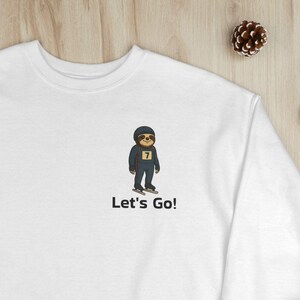 May include: White sweatshirt with a crew neck. The design features a cartoon sloth wearing sunglasses and a jersey with the number 7. The text "Let's Go!" is printed below the sloth. The sweatshirt is displayed on a plaid blanket with pine cones and Christmas decorations.