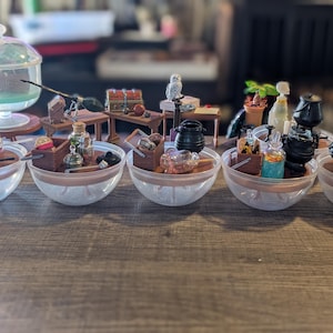 May include: A collection of miniature fantasy-themed dioramas featuring small plastic cauldrons, glass-like potion bottles, wooden crates, and tiny accessories arranged in clear plastic bowls and on small brown tables.