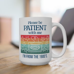 Puede incluir: Taza de cerámica blanca con un diseño retro que presenta una cinta de casete y estrellas. El texto en la taza dice "Please be PATIENT with me I'M FROM THE 1900'S."