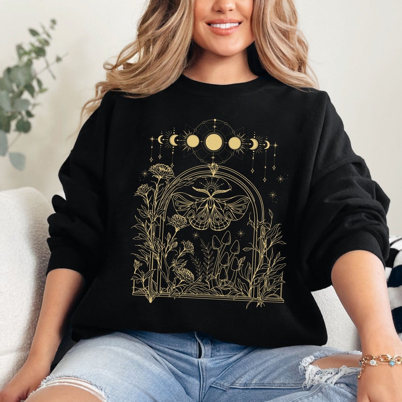 May include: Black sweatshirt with a gold graphic design of a moth, mushrooms, and flowers in a moonlit forest. The design includes a crescent moon, full moon, and stars.