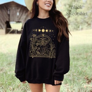 May include: Black sweatshirt with a gold graphic of a moth, flowers, and mushrooms under a crescent moon and stars. The text "Black Sweatshirt" is in the top right corner of the image.