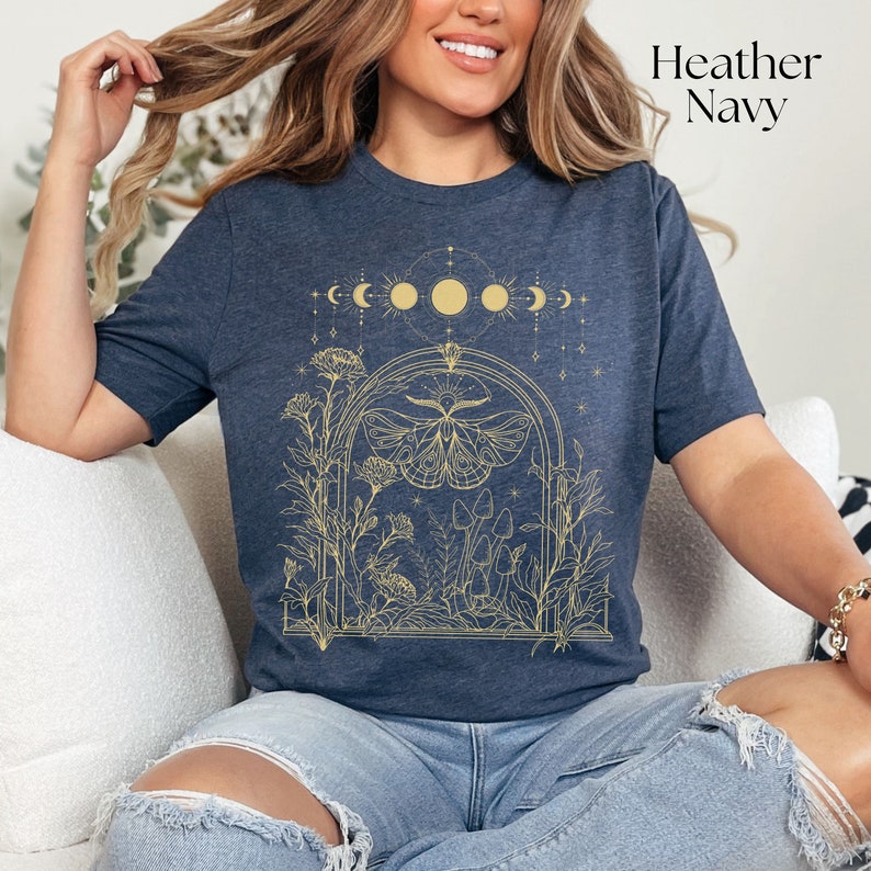 May include: A heather navy blue t-shirt with a gold outline graphic of a moth, mushrooms, and flowers in a moon phase arch. The text "Heather Navy" is in the top right corner of the image.