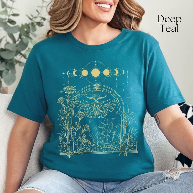 May include: A teal blue t-shirt with a gold outline design of a moth, mushrooms, and flowers in a window frame. The design also includes a crescent moon, sun, and stars.