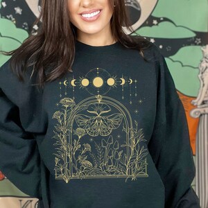 May include: A black sweatshirt with a gold graphic design featuring a moth, mushrooms, and a crescent moon with stars. The design is framed by a gold arch.