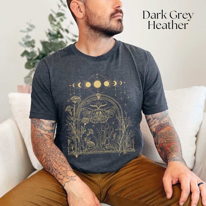 May include: A dark grey heather t-shirt with a gold graphic of a moth, flowers, and mushrooms. The graphic also includes a crescent moon and full moon design.