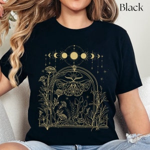 May include: Black t-shirt with a gold outline design of a moth, mushrooms, and a crescent moon with stars. The design is in a window-like frame with floral accents.