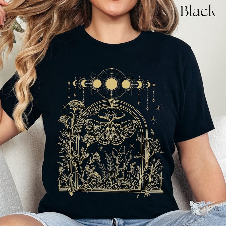 May include: Black t-shirt with a gold outline design of a moth, mushrooms, and a crescent moon with stars. The design is in a window-like frame with floral accents.