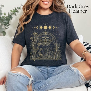 May include: Dark grey heather t-shirt with a gold outline of a moth, mushrooms, and flowers in a moonlit archway. The text "Dark Grey Heather" is in the top right corner of the image.