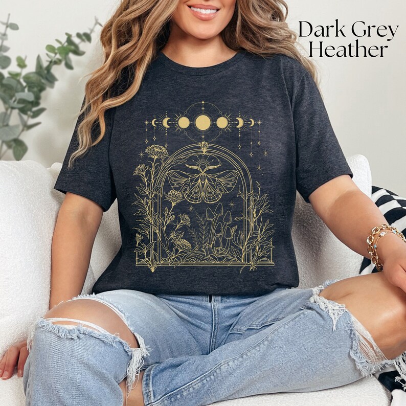 May include: Dark grey heather t-shirt with a gold outline of a moth, mushrooms, and flowers in a moonlit archway. The text "Dark Grey Heather" is in the top right corner of the image.