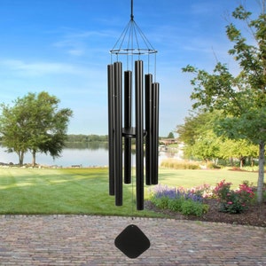 May include: Black wind chime with a diamond-shaped clapper hanging from a black metal frame. The chime is made of 8 tubes of varying lengths. The chime is hanging in a garden setting with a lake in the background.