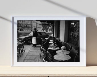 May include: Black and white photograph of a Parisian cafe scene. Two people are seated at a table outside, while a waiter stands nearby. A car is parked in the background.