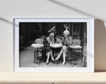 May include: Black and white photograph of two women sitting at a cafe table, drinking coffee. The cafe is outdoors and has a sign that says "Telephone".
