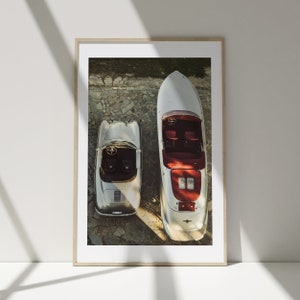 May include: A silver Porsche 356 Speedster and a silver speedboat are parked side-by-side on a cobbled driveway. Both vehicles are viewed from above.