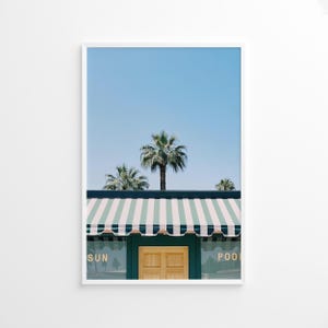 May include: Framed print featuring a sunny scene with palm trees against a clear blue sky. A building with a green and white striped awning and a wooden door is visible. The words "SUN" and "POOL" are on the glass.