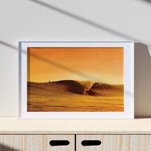 May include: A framed photograph of a large wave breaking in the ocean at sunset. The wave is a golden yellow color, and the sky is a deep orange. The image is framed in a white frame.