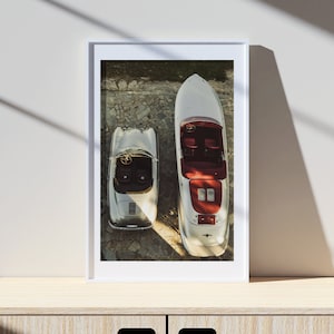 May include: A framed photograph of a silver car and a silver speedboat, both viewed from above. The car and boat are parked on a stone surface. The car has a red interior and the boat has a red interior and a red stripe.