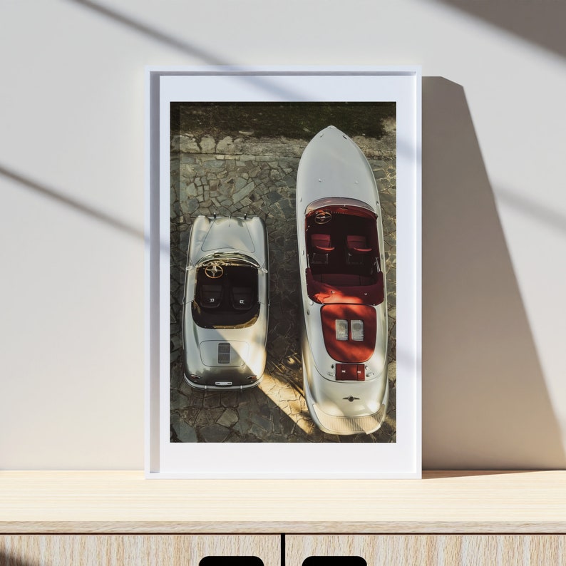 May include: A framed photograph of a silver car and a silver speedboat, both viewed from above. The car and boat are parked on a stone surface. The car has a red interior and the boat has a red interior and a red stripe.