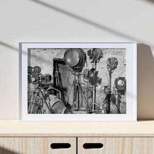 May include: A black and white photo of a film set with a vintage movie camera and several spotlights on tripods.