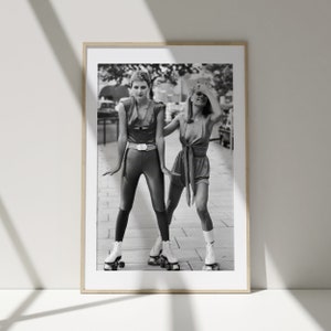 Pu&ograve; includere: Due donne con abiti in stile anni '70 stanno pattinando su una strada cittadina. Una donna indossa una tuta nera con una cintura e l'altra indossa un vestito corto con un nodo. Entrambe le donne indossano pattini bianchi.