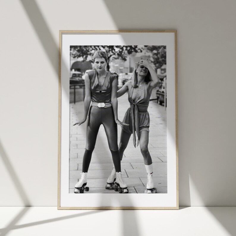 Pu&ograve; includere: Due donne con abiti in stile anni '70 stanno pattinando su una strada cittadina. Una donna indossa una tuta nera con una cintura e l'altra indossa un vestito corto con un nodo. Entrambe le donne indossano pattini bianchi.
